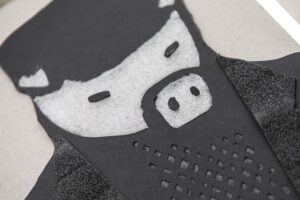 Zbliżenie na tyflografikę do obrazu „Maus 5” (2001, olej na płótnie) Wilhelma Sasnala. Twarz świni. Na głowie ma kaszkiet, ubrana jest w kraciastą koszulę i czarną marynarkę. Świnia namalowana przez Sasnala przypomina człowieka. 