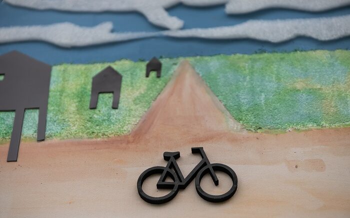 Zbliżenie na część tyflografiki „Bez tytułu”. Wypukły rower, za nim ścieżka, dookoła zieleń. Po lewej stronie tyflografiki trzy czarne wieże strażnicze. Za horyzontem niebo.