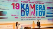 Dwie osoby stoją na scenie, za nimi grafika zapraszająca na festiwal filmowy